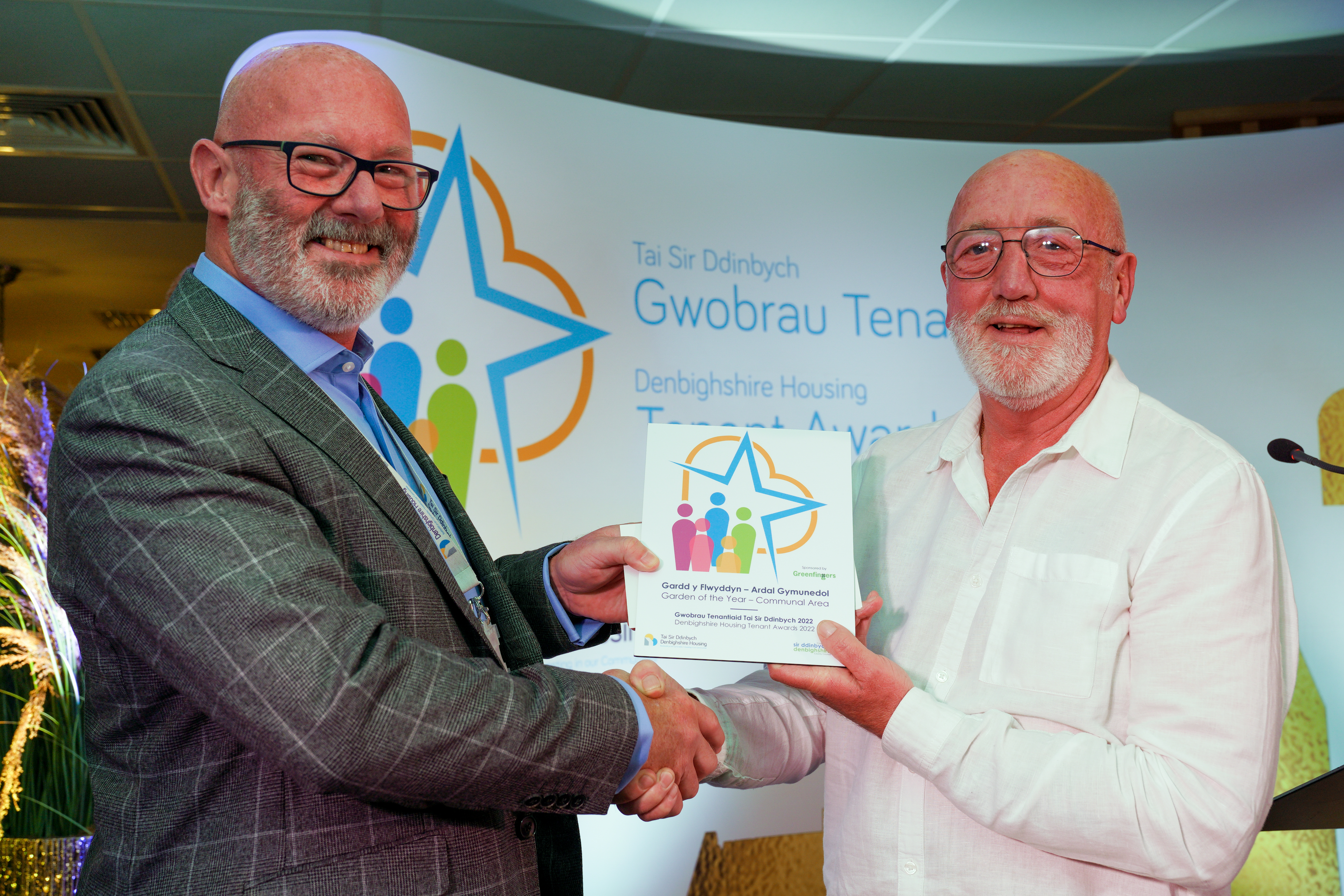 Two men stand together at an award event, one wears a grey suit and is presenting a colourful glass award to the man in a white shirt.