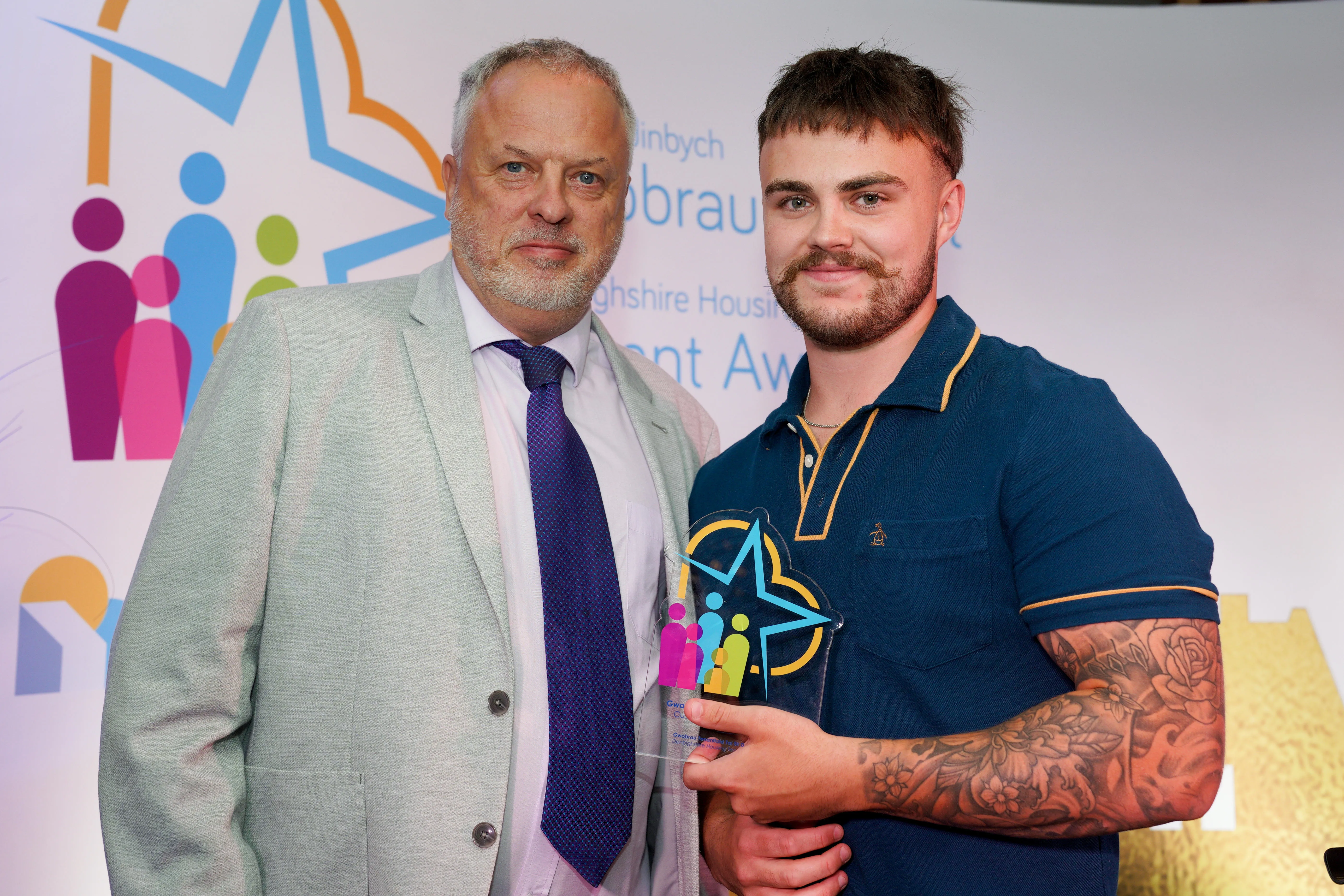 Two men stand together at an award event, with the younger man holding a colourful trophy.