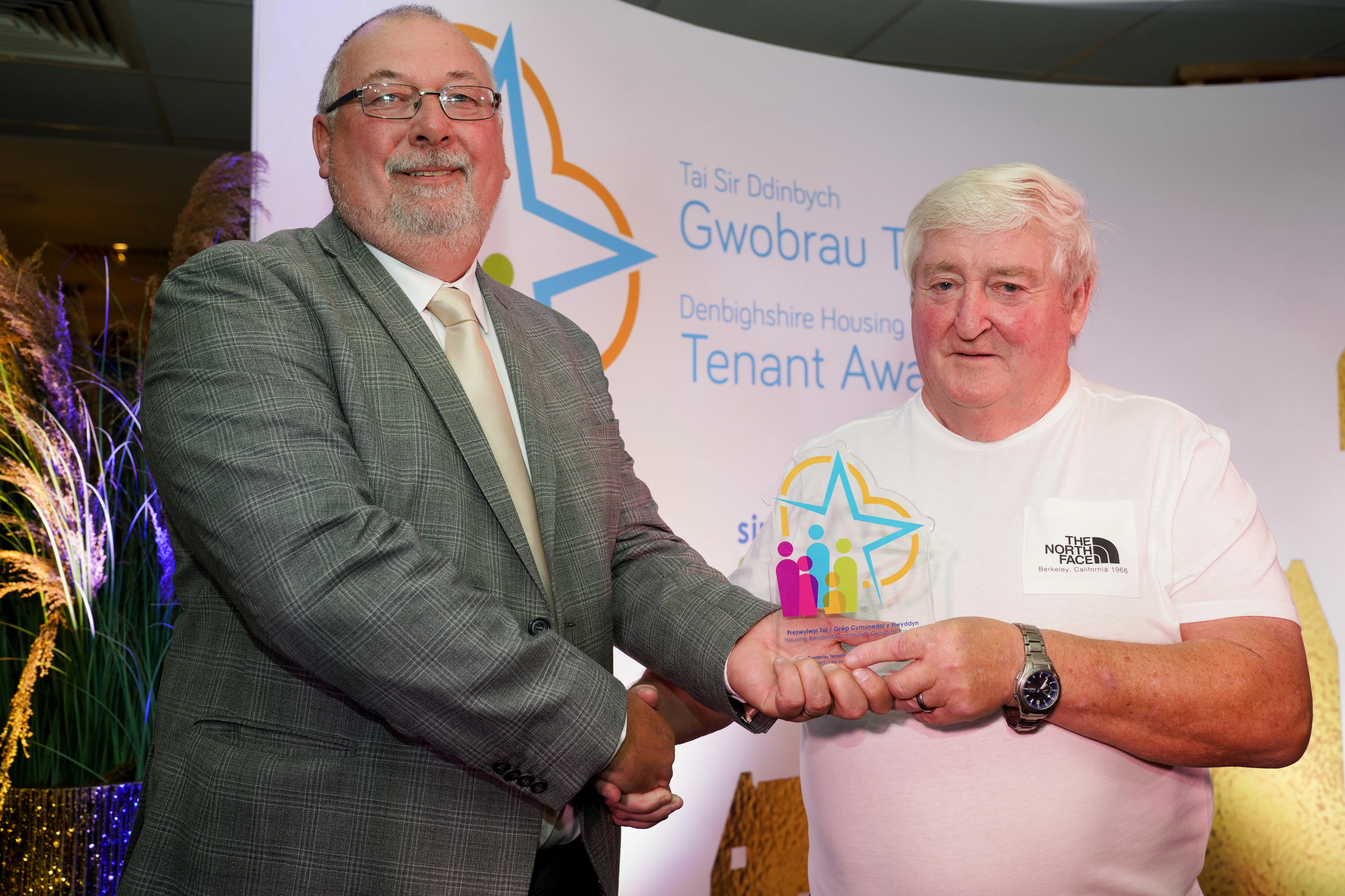 Two men stand together at an award event. A man in a grey suit hands a colourful trophy to a man wearing a white t-shirt.