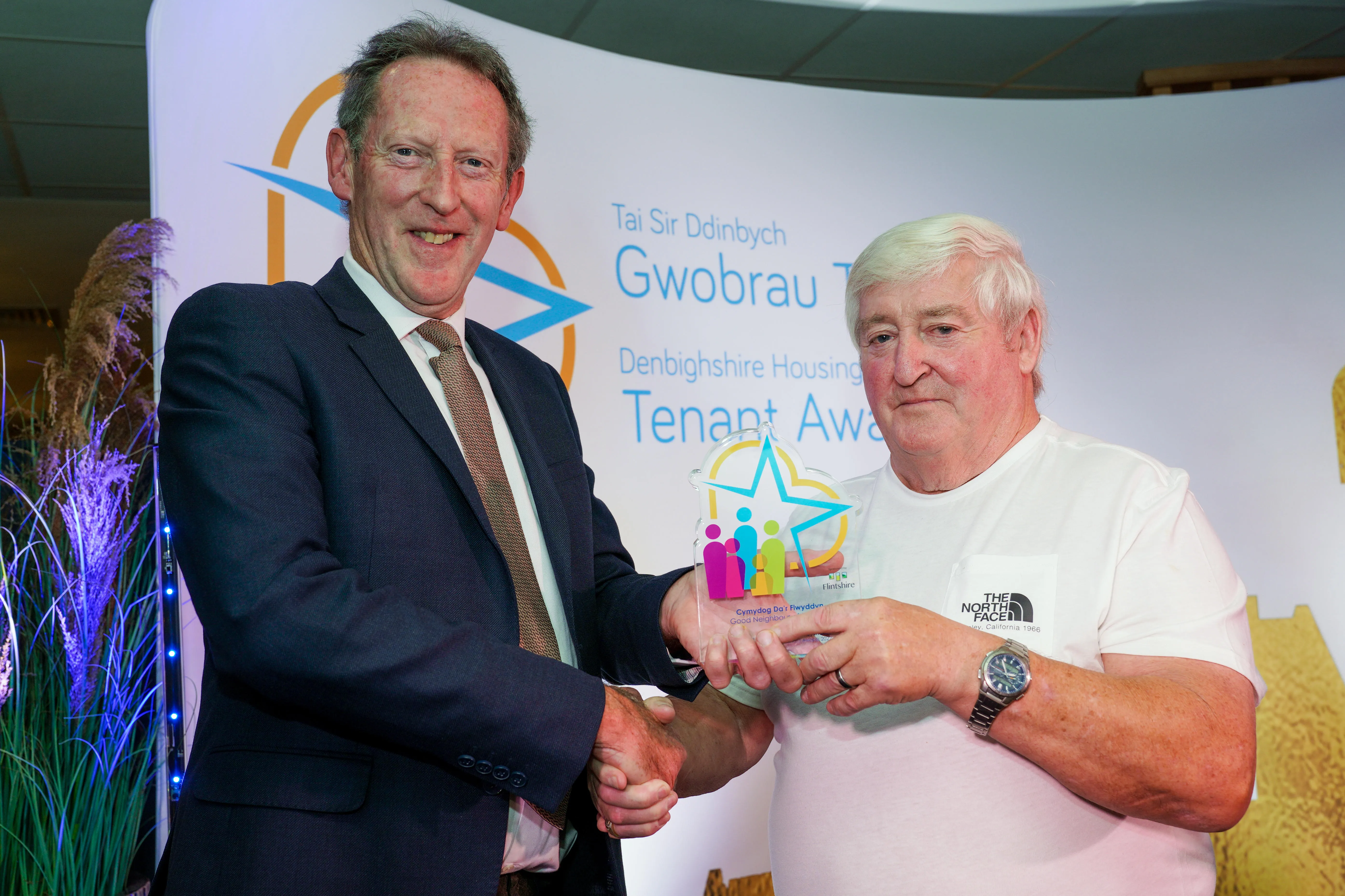 Two men stand together at an award event, with the man in a blue suit handing a colourful award to the man wearing a white top.