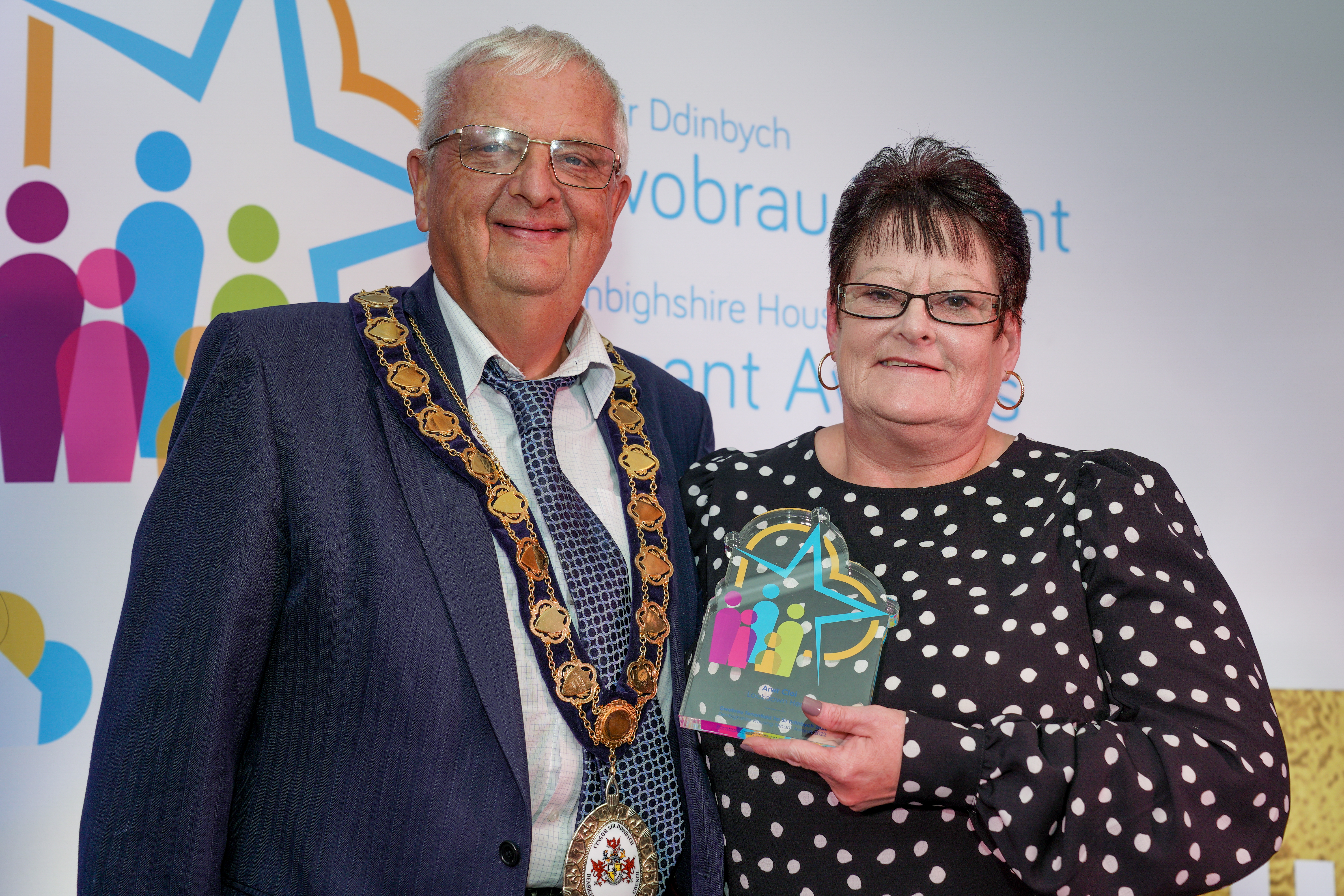 A man and woman stand together at an award event. The man is wearing civic regalia to present a glass award to the woman.