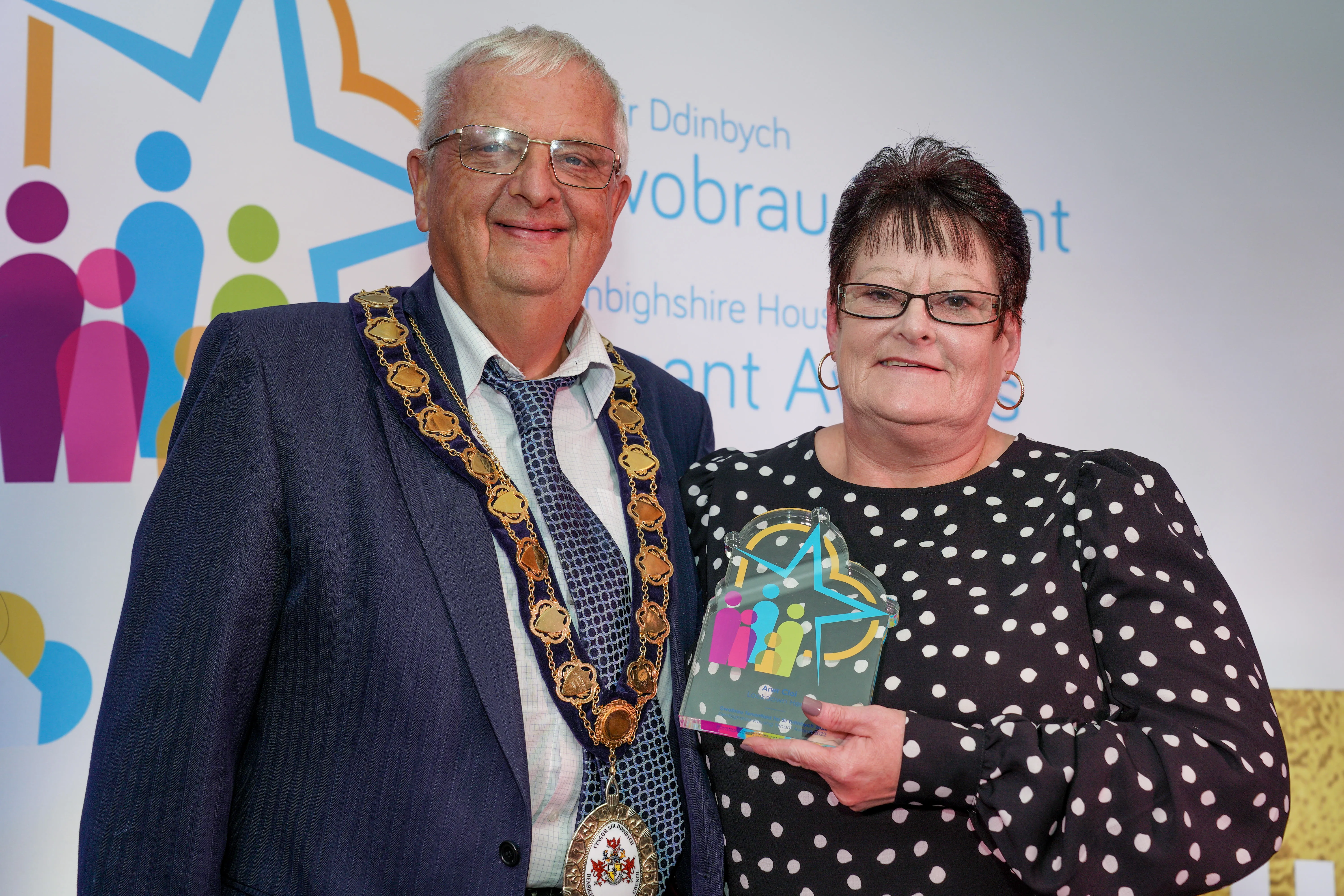 A man and woman stand together at an award event. The man is wearing civic regalia to present a glass award to the woman.